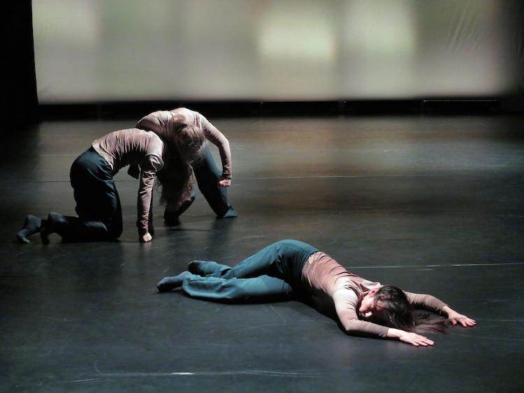 trio of dancers in triangle formation, one lying on floor as the other two huddled behind her are conjoined and rising from a lower position
